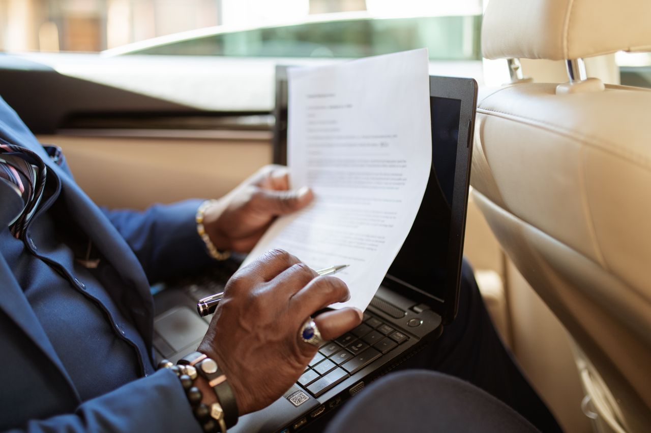 Offerings A businessman reviews documents on a laptop while traveling in the backseat of a car.